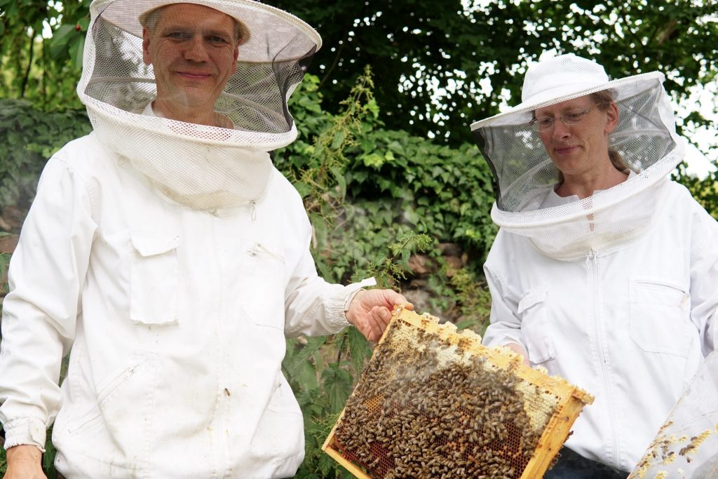 Ein Mann und eine Frau stehen in Imkerkleidung vor einer Wand.